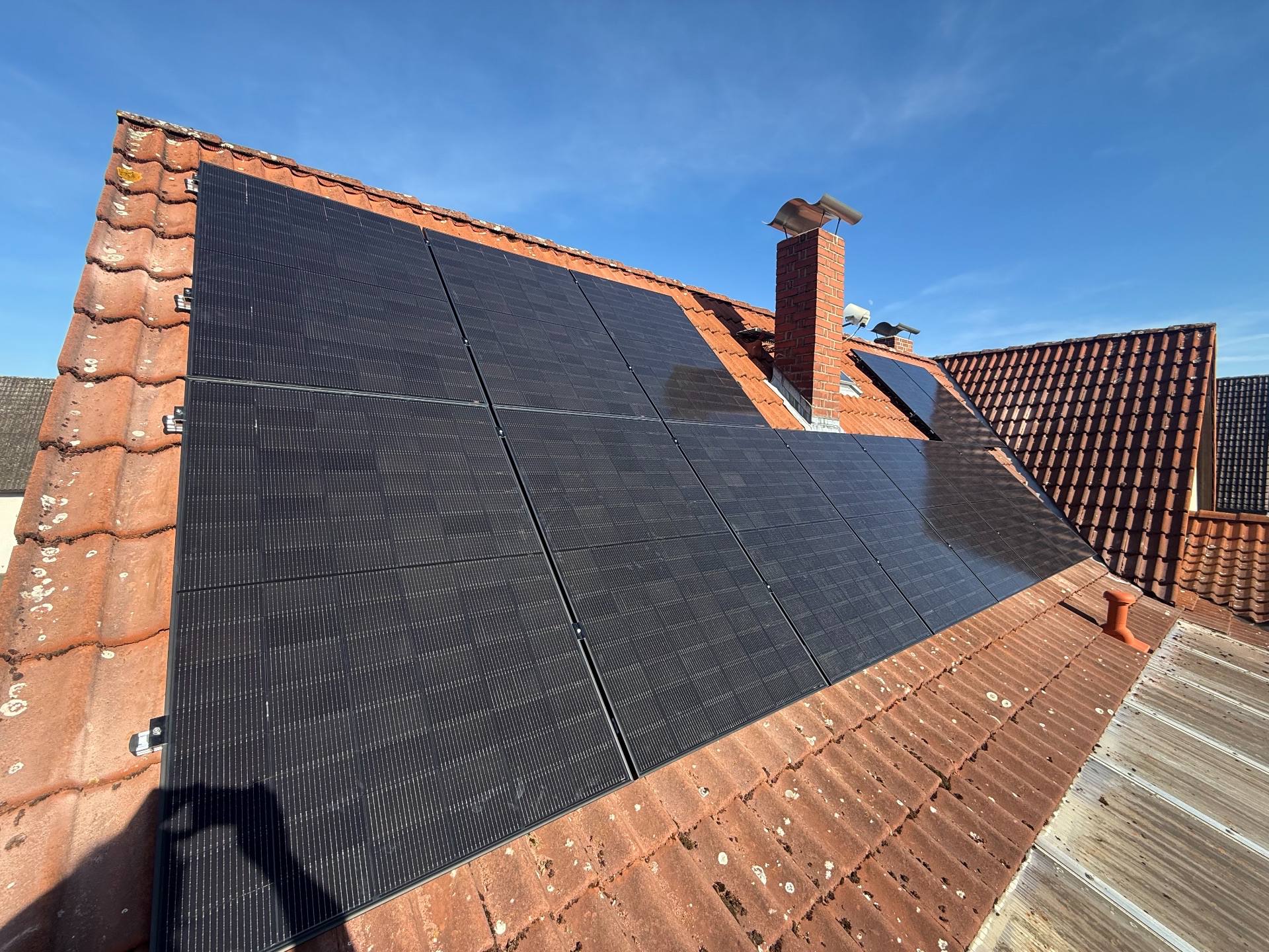 Schwarze Solarmodule aus der N&auml;he auf einem roten Ziegeldach, daneben ein Backstein-Schornstein